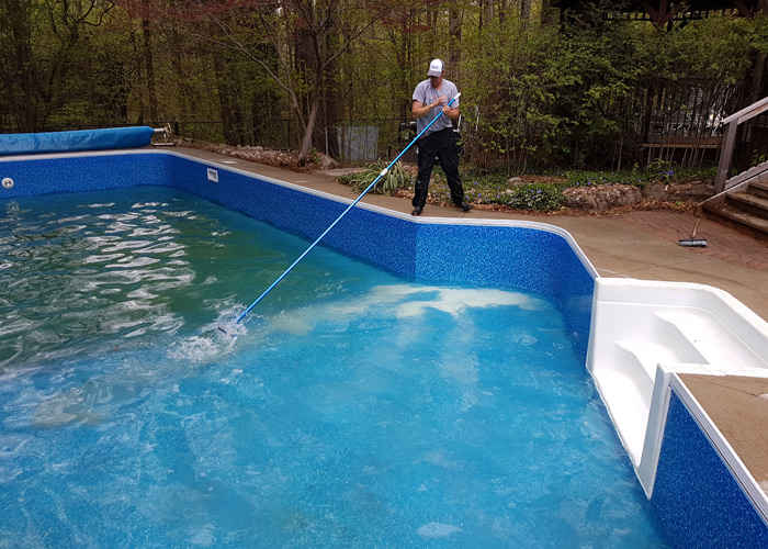 Person cleaning a backyard in-ground pool using a long pool skimmer pole. The pool has a blue liner, white steps, and is surrounded by trees and a garden. A pool cover is partially visible on the left side.