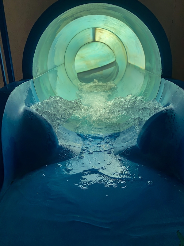 Interior of a clear water slide with rushing water and a light at the end.