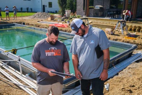 Two men standing by a pool under installation look down at a document together.