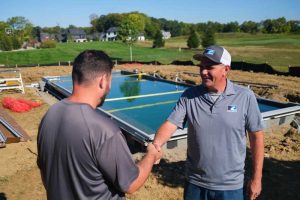 Two men shake hands standing in front of a pool being installed.