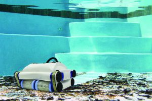 An underwater view of a robotic pool cleaner removing debris from the pool floor near the steps, with clear water and tiled walls in the background.