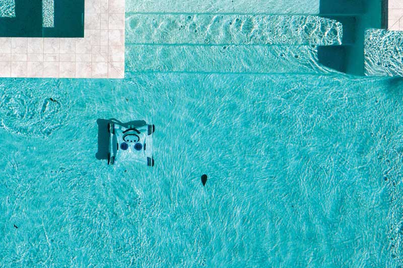 An overhead view of a swimming pool with crystal-clear water, featuring a robotic pool cleaner in operation near the tiled pool edge.