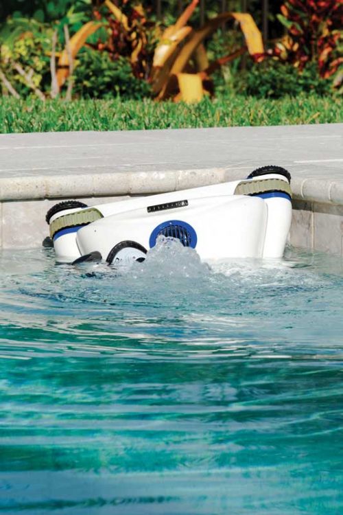 A robotic pool cleaner operating in a pool corner, creating water turbulence as it scrubs near the tiled edge, with landscaping visible in the background.