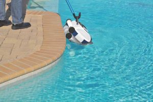 A person standing on the pool deck uses a pole to lift a robotic pool cleaner out of the water, with the clean blue pool surface in the background.