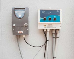 A close-up of a pool automation system mounted on a wall, featuring a control panel with labeled buttons and an adjacent AC transformer unit.