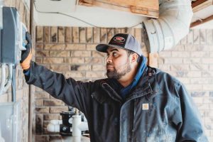 A technician in a jacket and cap works on a pool automation control system mounted on a brick wall, with additional pool equipment visible in the background.