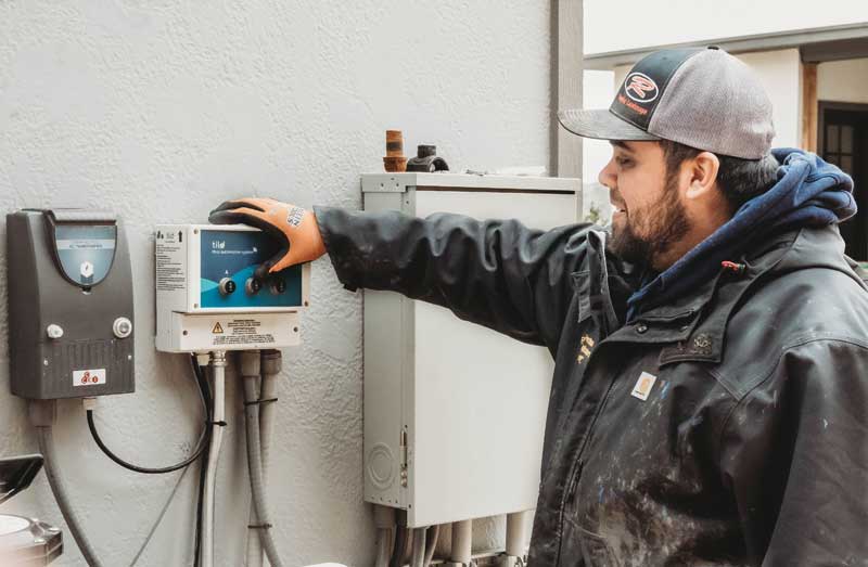 A technician in a cap and jacket adjusts a pool automation control panel mounted on a wall, with additional equipment visible nearby.