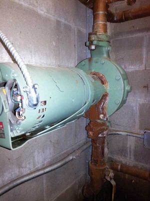 A close-up view of an industrial water pump connected to corroded pipes, installed on a concrete wall in a mechanical room.