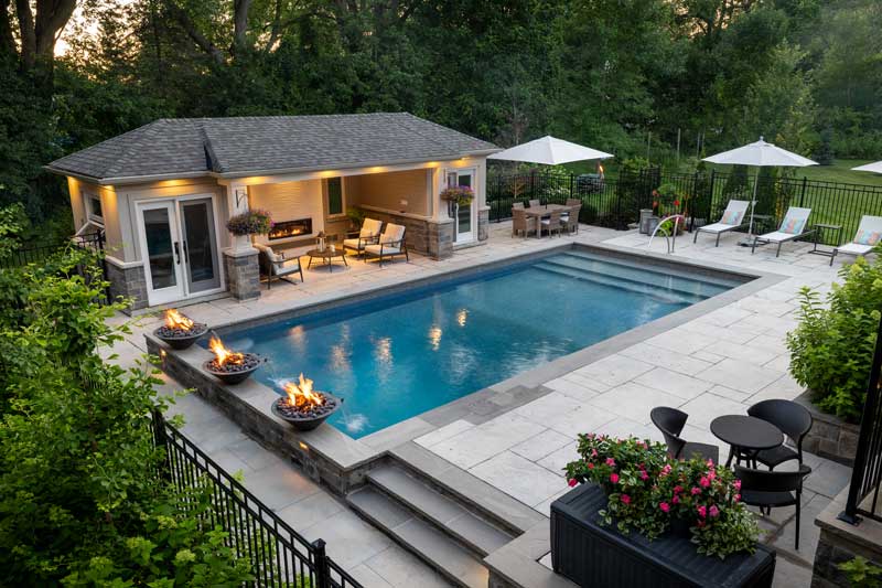 A luxurious backyard featuring a swimming pool surrounded by stone tiles, fire bowls, a pool house, lounge chairs, and lush greenery in the evening light.