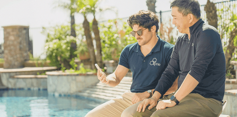 Two men seated poolside, one demonstrating a smartphone app, showcasing simple pool automation technology amidst a vibrant, sunny outdoor setting.