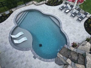 Aerial view of a uniquely-shaped backyard pool surrounded by a clean paver deck, lounge chairs, and landscaped greenery, creating a luxurious outdoor space.