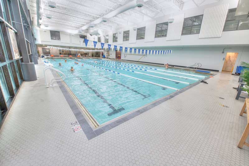 A spacious indoor swimming pool with marked lanes, blue-and-white flags hanging above, and swimmers enjoying the facility, surrounded by tiled flooring and large windows.