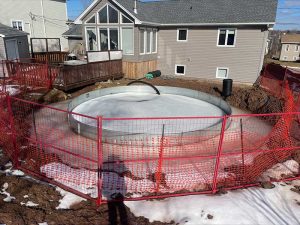 Partially constructed swimming pool surrounded by red safety fencing in a residential backyard, with snow patches and a nearby home in view.