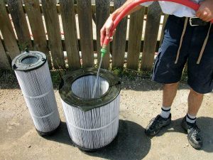 A person rinsing large pool filter cartridges with a hose outdoors near a wooden fence on a sunny day.