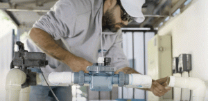 A technician in a white cap and sunglasses installing a pool automation device, surrounded by white PVC piping in a maintenance area. 