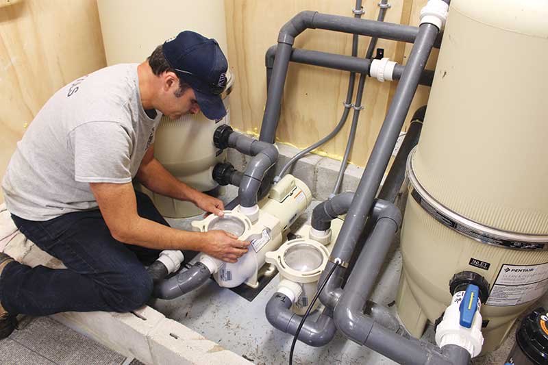 A pool technician inspecting and cleaning a pool pump and plumbing system in a dedicated indoor equipment room.