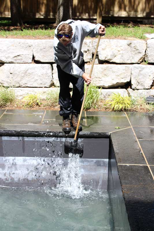 A worker wearing sunglasses and a hoodie uses a brush on a pole to clean a swimming pool, with stone landscaping in the background.