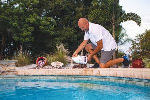 A person pouring liquid pool treatment from a bottle into a backyard pool surrounded by plants and landscaping.