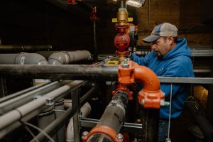 A technician wearing a blue hoodie and cap works on a complex system of pipes and valves in a mechanical room, with dim lighting and industrial equipment surrounding him.