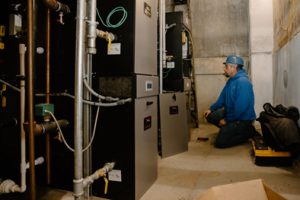 A technician in a blue hoodie and hard hat kneels in a mechanical room, working on a row of heating systems with visible pipes and wiring.