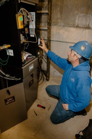 A technician wearing a blue hoodie and hard hat works on a heating system, using diagnostic tools and wiring equipment in an industrial setting.