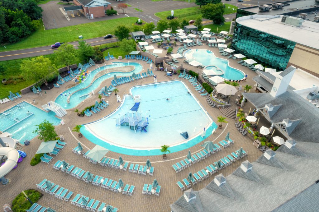 An aerial view of a large outdoor water park featuring multiple pools, a lazy river, water slides, and lounge chairs arranged around the deck, with greenery and buildings in the background.