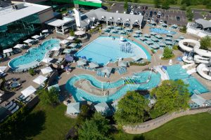 An aerial view of a sprawling outdoor aquatic facility featuring multiple pools, a lazy river, water slides, lounge areas, and shaded seating, with adjacent parking and greenery.