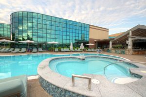 A modern outdoor pool area with a spa-like design, surrounded by lounge chairs and umbrellas, set against a building with reflective glass windows.