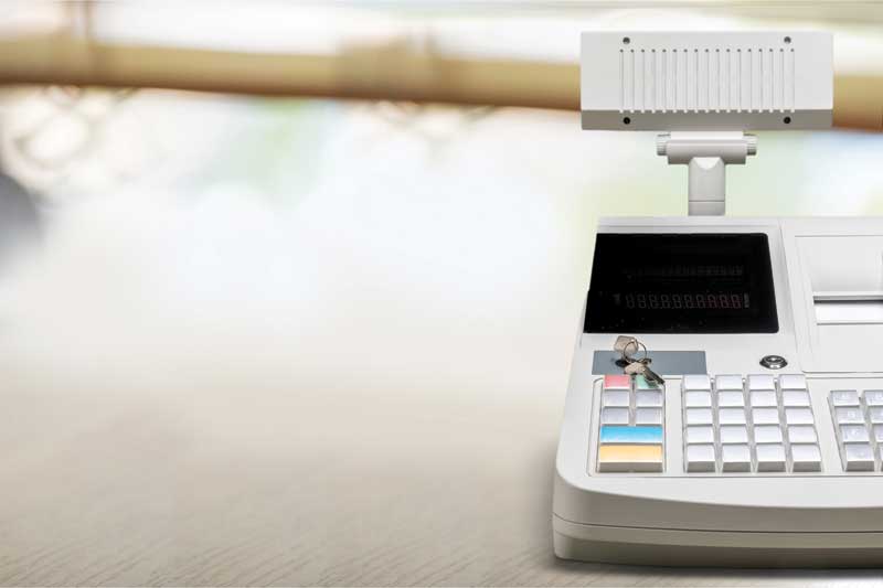 A close-up of a cash register with a display screen and keypad on a wooden counter, set against a blurred background.