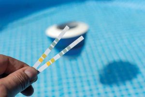 A hand holding two pool test strips against the background of a blue swimming pool, with a floating pool skimmer visible in the water.