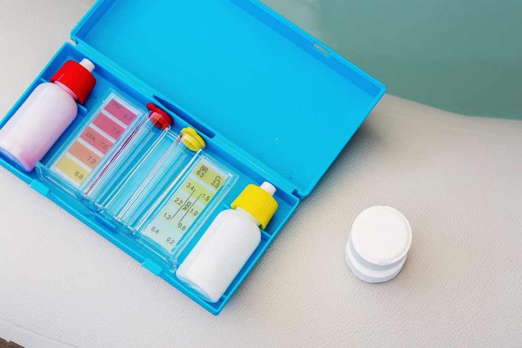 A pool water testing kit with pH, chlorine, and bromide test components in a blue case, alongside a white pool chemical tablet.