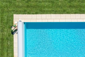 An aerial view of a worker in green uniform servicing a residential swimming pool, surrounded by a neatly trimmed lawn and clean blue water.