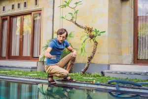 A man crouches by a swimming pool holding a water test kit, checking chemical levels, with a garden and house in the background.