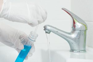 A close-up of hands in gloves using a pipette to add blue liquid into a test tube next to a running water tap.