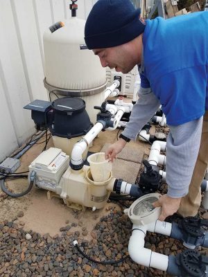 A technician inspecting a pool pump system outdoors, wearing a blue uniform and beanie, surrounded by pipes and equipment.