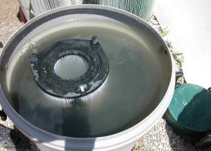 A large pool filter soaking in a container of dirty water during maintenance, surrounded by equipment outdoors.