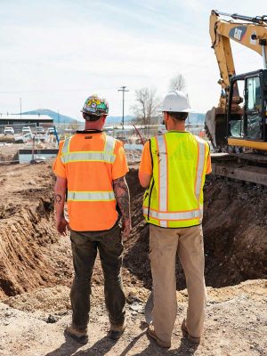 Two construction workers in reflective vests and hard hats stand at a worksite, facing an excavator and observing the progress of excavation.