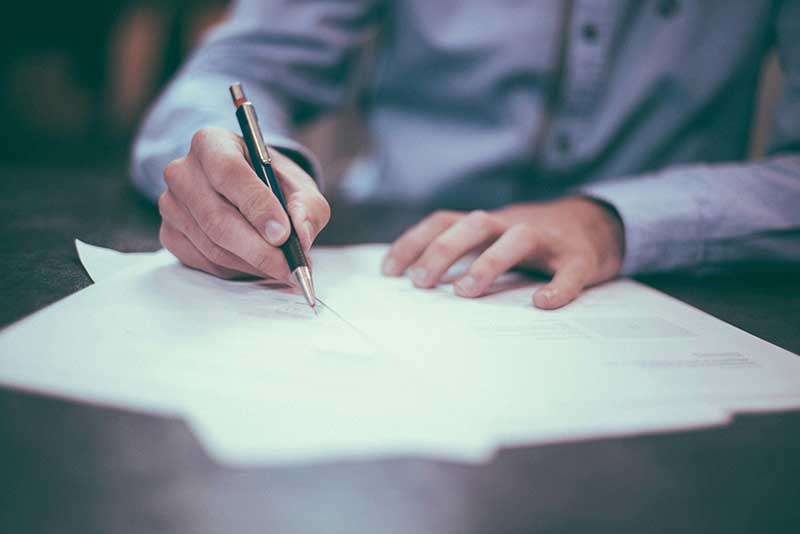 A close-up of a person in a dress shirt holding a pen and writing on paper documents spread across a desk or table.