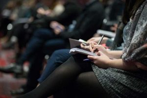 Audience members seated, taking notes during a conference or lecture, highlighting active participation and engagement in a professional or educational setting.