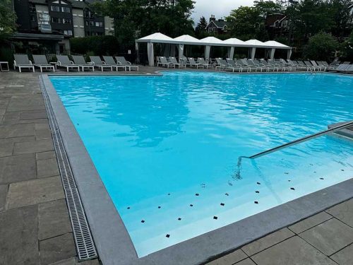 Outdoor swimming pool with clear blue water, surrounded by a paved deck, lounge chairs, and white cabanas, set against a backdrop of greenery and buildings.