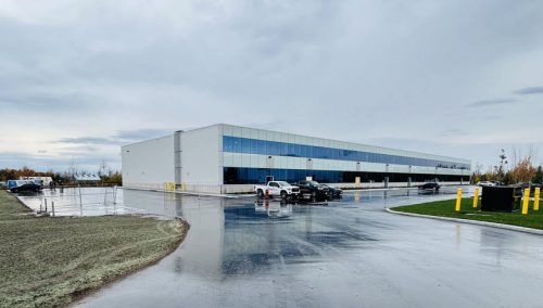 A modern, two-story office and warehouse facility with large glass windows, surrounded by a wet pavement parking lot under an overcast sky. The building is on a spacious lot with grassy areas and scattered trees in the background.