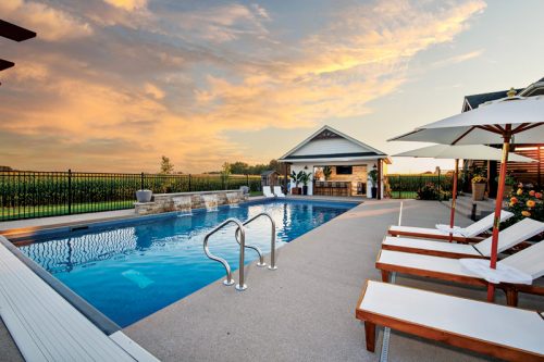 A serene pool area features sparkling water surrounded by lounge chairs. An outdoor kitchen is visible in the background, complemented by the colorful sunset sky. A black fence borders the area, with lush greenery beyond.