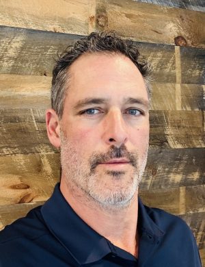 A middle-aged man with short, slightly wavy gray hair and a well-groomed beard is looking directly at the camera. He is wearing a navy blue polo shirt. The background features a textured wooden wall with visible knots and varying wood tones, creating a warm, natural atmosphere.