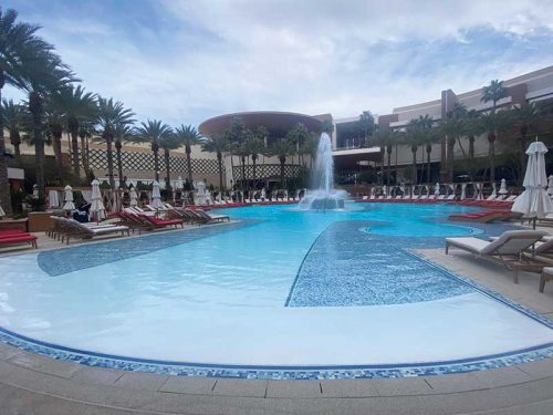 Luxurious resort-style pool with a central fountain, surrounded by palm trees, red and white lounge chairs, and umbrellas, set against a backdrop of elegant architecture and a partly cloudy sky.