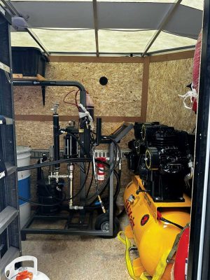 Interior of a compact equipment shed featuring a yellow air compressor, plumbing tools, hoses, a fire extinguisher, and an organized workspace with plywood walls and a translucent roof.