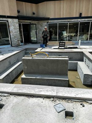 Construction site of a residential or commercial pool under development, featuring unfinished concrete structures, a worker in the background, and modern sliding glass doors with stone and wood-paneled exterior walls.