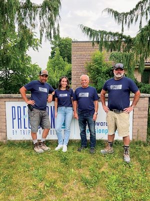 Group photo of four people wearing matching 'Precision' branded shirts, standing in front of a brick wall with a company sign, surrounded by greenery.