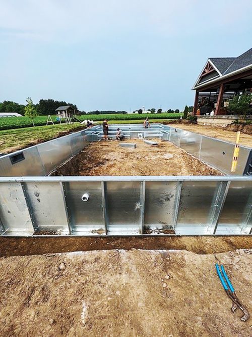 Four workers are constructing a rectangular pool, surrounded by dirt and gravel. The pool walls are made of shiny metal sheets, and nearby, there is a house with a porch, along with a swing set in the background. The sky is mostly clear, indicating a sunny day.