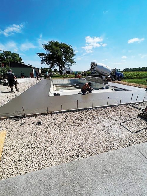 Workers are pouring concrete for a construction foundation in a rectangular shape. The site is set against a clear blue sky with scattered clouds, and a truck with a cement mixer is visible in the background. A large tree and some buildings are also seen in the area, and gravel is laid down on the ground.