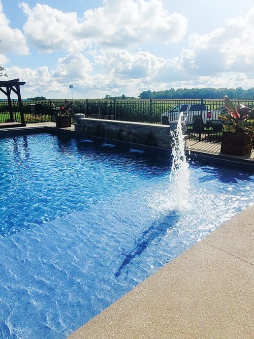 A sparkling blue swimming pool features a central fountain that sprays water into the air. Lush plants and a wooden structure are visible beside the pool, with a fenced area in the background. The scene is illuminated by sunlight and dotted with fluffy white clouds.